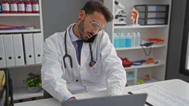 Klinikteki genç İspanyol adam steteskop takıyor. Laptop ve telefon kullanıyor. Modern bir hastane işyeri sergiliyor..