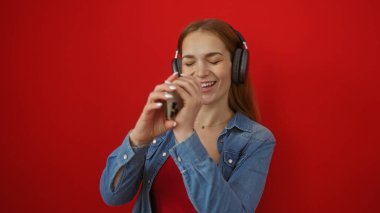 Kulaklıklı müzikten hoşlanan bir kadın canlı, modern atmosferde neşe ve tutku taşıyan canlı kırmızı bir arkaplan karşısında telefona gülümseyip şarkı söylüyor..