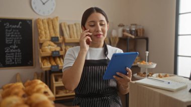 Genç, İspanyol, tablet kullanan ve fırında telefonla konuşan, arka planda ekmek olan, sipariş detaylarını kontrol eden bir kadın.