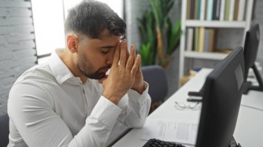Beyaz gömlekli genç adam elleri ofis masasında oturuyor. Etrafı bilgisayar ve evrak işleriyle çevrili. Profesyonel bir ortamda stres taşıyor..