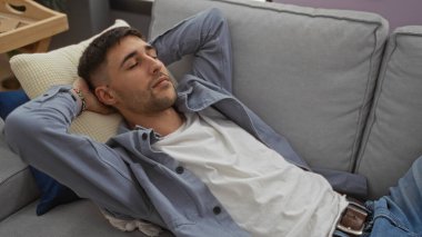 Kapalı bir koltukta, kolları kafasının arkasında rahat bir şekilde, rahat ve huzurlu bir şekilde, modern bir ev ortamında rahat bir şekilde uzanan genç bir adam..