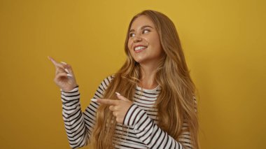 Çekici sarışın kadın gülümsüyor ve çizgili gömleğiyle izole edilmiş sarı duvar arkasını işaret ediyor.