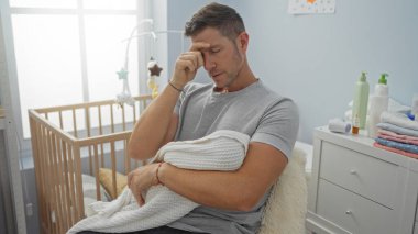 Sıcak yatak odasında bebeği tutan genç bir adam, yorgun bir şekilde oturuyor. Huzurlu bir atmosferi olan, beşikteki hafifçe aydınlatılmış odada..