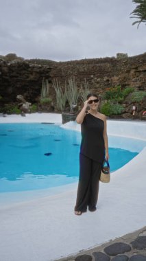 Lanzarote 'de mavi havuzun yanında duran siyah elbiseli ve güneş gözlüklü Jameos del agua' da sepetli bir kadın.