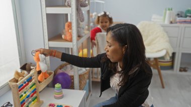 Bebek odasında oyuncak ile oynayan bir kadın etrafı doldurulmuş hayvanlar, abaküs ve canlı dekorlarla çevrili, aile bağını ve öğrenme ortamını sembolize ediyor..