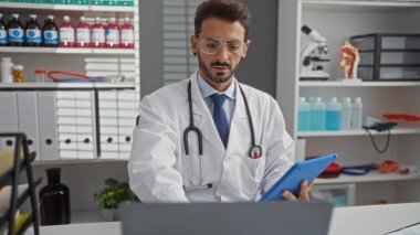 Klinikte gözlüklü ve laboratuvar önlüklü genç İspanyol erkek doktor, profesyonel bir sağlık ortamında tıbbi dosyalar ve ekipmanlarla çevrili bir tableti inceliyor..