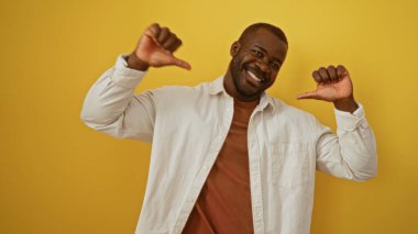 Genç adam kendinden emin bir şekilde kendini işaret ederek gülümsüyor. Parlak sarı arka plan üzerinde başparmakları var.