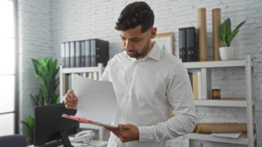 Modern bir ofiste doküman okuyan bir adam, raflar, bitkiler ve ciltlerle süslü bir iç mekan, profesyonel ve odaklanmış bir iş ortamı yakalıyor..
