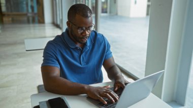 Genç, Afrikalı, Amerikalı, modern ofis ortamında çalışan gözlüklü ve mavi gömlekli bir adam.