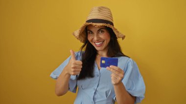 Baş parmağıyla gülümseyen ve izole edilmiş sarı arka planda hasır şapka takan bir kadın.