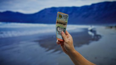 Avustralya 'nın deniz kenarındaki banknotlarıyla el ele tutuşmuş. Sahilde manzaralı bir yerde bir adamla iş bağlamında para değişimi tasvir ediyor..