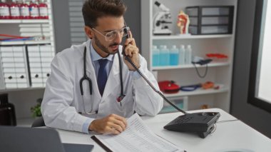 Klinikteki genç İspanyol erkek doktor gözlük ve laboratuvar önlüğü takıyor, telefonla konuşuyor, belgeleri inceliyor, tıbbi ekipmanlar ve parlak hastane odasındaki raflarla çevrili.