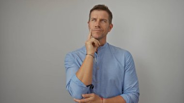 Mavi gömlekli genç adam özenle beyaz bir arka plana karşı poz vererek izole edilmiş bir stüdyo ortamında tefekkür ve güven hissi veriyor..