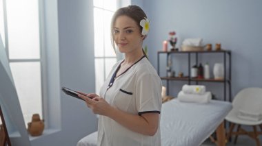 Tablet tutan, üniformalı, saçları çiçekli, modern kaplıca salonunda duran bir kadın.