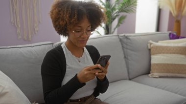 Akıllı telefon kullanırken gülümseyen bir kadın, mor duvarları ve içinde bitki ve yastıkların bulunduğu modern dekorasyonu olan rahat oturma odasında rahat gri koltukta oturur..