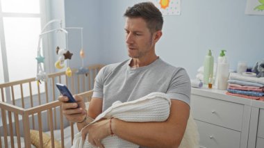 Bebeği yatak odasında tutarken akıllı telefon kullanan bir adam, bebeğin temel ihtiyaçları, beşik ve yumuşak aydınlatma ile çevrili, sakin ve şefkatli bir atmosfer yaratıyor..