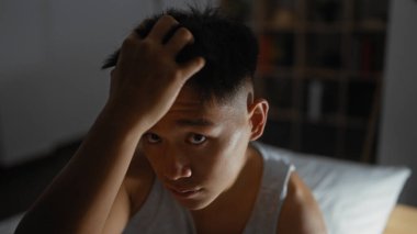 Genç adam kapalı bir odada oturuyor, saçlarına dokunurken düşünceli görünüyor, loş ışıklı bir odada yüzünü aydınlatan yumuşak gölgelerle..