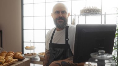 Sakallı ve dövmeli İspanyol bir adam, fırının arkasında duruyor, önlük giyiyor ve gülümsüyor, kruvasan, hamur işleri ve fırında pişirilmiş mallarla.