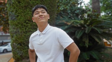 Beyaz gömlekli genç adam şehir parkında güvenle duruyor. Arka planda yeşillik, modern şehir yaşam tarzını gözler önüne seriyor.