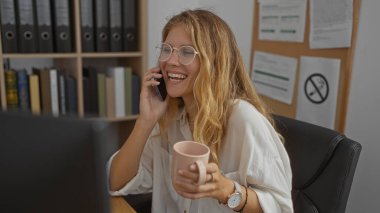 Ofiste kahve fincanı tutan ve arka planda kitap rafları olan genç bir kadın telefonda konuşuyor.