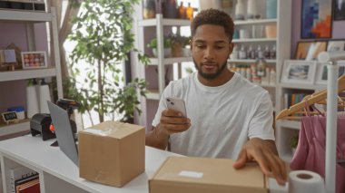 Evin dekorasyon dükkanında çalışan genç bir adam akıllı telefon kullanarak kapalı mekan bitkileri ve dekorasyonlarla çevrili bir tezgahta paketleri yönetiyor.