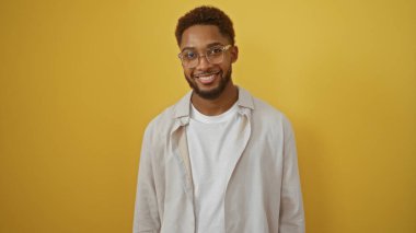 İzole edilmiş sarı bir arka planda gülümseyen yakışıklı genç adam, günlük kıyafetler ve gözlük takıyor.