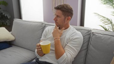 Düşünceli bir suratı olan genç bir adam gri bir kanepede oturan sarı bir bardağı tutuyor rahat bir oturma odasında yumuşak ışık ve doğal bir dekorla..