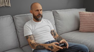 Sakallı ve dövmeli İspanyol bir adam oturma odasındaki kanepede video oyunu oynuyor ve yüzünde odaklanmış bir ifade var.