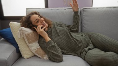 Kadın evdeki kanepede dinleniyor, telefonda gülümsüyor, rahat kıyafetler giyiyor, rahat bir apartman dairesinde minderlerle çevrili, sıcak, davetkar bir atmosfer sergiliyor..