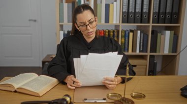 Kadın yargıç bir ofis salonunda oturuyor, esmer kadın endişeli bir ifadeyle belgeler okuyor, masasında hukuk kitapları ve adalet terazileriyle çevrili.