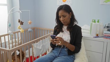 Yatak odasında oturan bir kadın beşiğin yanında telefon kullanıyor ve bebek yukarıda asılı duruyor, sevgi ve bağlılık ile modern kapalı alan aile ortamını sergiliyor.