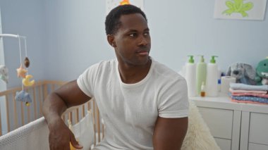 Yatak odasında oturan genç adam, beşikte düşünceli görünüyor, etrafı bebek eşyalarıyla çevrili, ilgili bir ev ortamı öneriyor..