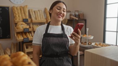 Fırında genç bir kadın gülümseyerek, içi taze ekmek ve hamur işleriyle dolu raflara bakıyor.