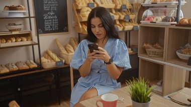 Kapalı bir pastanede, rafları ekmek ve hamur işleriyle dolu akıllı telefon kullanan genç bir kadın kahve fincanıyla bir masada oturuyor.