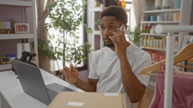 Ev dekorasyonu mağazasında çalışan genç bir adam, telefonda konuşurken arka planda dekorasyon rafları olan bir dizüstü bilgisayar kullanıyor..