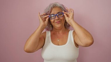 Gri saçlı ve gözlüklü orta yaşlı İspanyol bir kadın izole edilmiş pembe bir arka plana gülümsüyor.