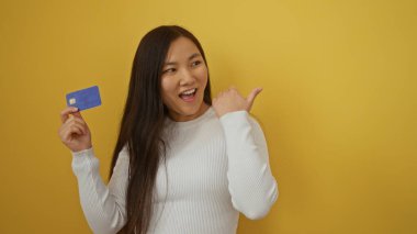 Elinde kredi kartıyla, hayat dolu sarı bir arkaplanı işaret eden bir kadın neşeli ve kendinden emin ifadesini vurguluyor..