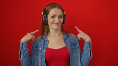 Kulaklık takan kadın, kot pantolon ve çekici ifade tarzını gösteren kırmızı arka plana karşı kendine güvenle işaret ediyor..