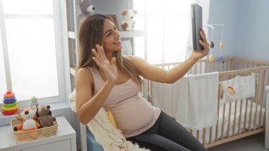 Beşik ve oyuncaklarla dolu rahat çocuk odasında tablete video izlerken el sallayan kadın, ev ortamındaki sıcaklığı ve bağlantıyı yakalıyor.