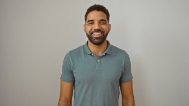 Genç İspanyol adam gülümsüyor, soyutlanmış beyaz bir arkaplanda dikiliyor, sıradan bir polo tişört giyiyor, çekici ve arkadaş canlısı bir tavır sergiliyor..