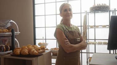 Fırında kolları çapraz duran, kahverengi önlüklü, etrafı hamur işleri, ekmek, pasta ve büyük pencereli fırın ürünleriyle çevrili bir kadın..