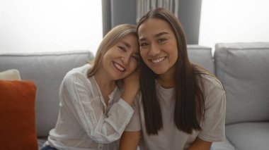 Anne ve kızı sıcak bir oturma odasında birlikte gülümsüyor, sevgi ve aile bağı sergiliyorlar..