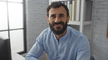 Sakallı yakışıklı bir adam ofiste güvenle gülümsüyor, açık mavi bir gömlek giyiyor ve arka planda rafları ve pencereleri olan bir masanın yanına oturuyor..