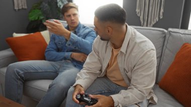 Erkekler bir dairede kanepede oturup oyun oynuyorlar, sohbet ediyorlar ve arka planda turuncu yastıklarla vakit geçiriyorlar..