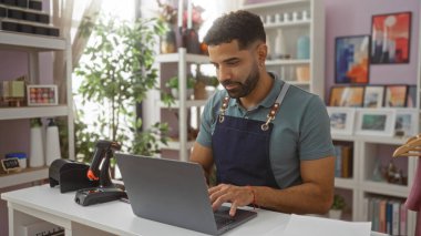 Dizüstü bilgisayarda çalışan genç bir adam dekorasyon rafları ve bitkilerle dolu bir mağazada çalışıyor.