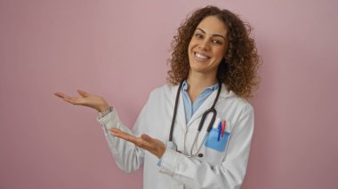 Steteskoplu beyaz önlüklü kadın doktor pembe arka planda gülümsüyor ve sunuyor. Profesyonel sağlık hizmetlerini güven ve arkadaşlıkla sunuyor..