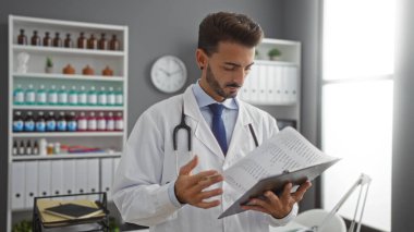 İspanyol erkek doktor, tıbbi malzemelerle çevrili iyi organize edilmiş bir klinikteki hasta dosyalarını analiz ediyor, profesyonelliğini gösteriyor ve işyerine odaklanıyor..