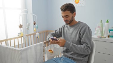 Yatak odasında, bebek beşiğinin yanında akıllı telefon kullanan genç bir adam ev içinde parlak bir beşik, rahatlamış bir iç mekanla ebeveynlik ve teknoloji bağlantısını öneriyor..