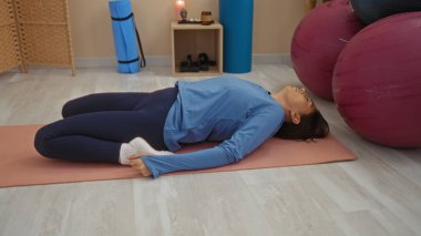 Spor merkezinde yoga yapan bir kadın paspasın üzerinde uzanmış kahraman pozu veriyor mavi spor kıyafetleri giyiyor ve rahatlama ve esneme için egzersiz ekipmanlarıyla çevrili.