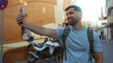 Arka planda motosikletle selfie çeken genç adam, sırt çantası takıyor, şehir ortamında samimi anlar yakalıyor, gençlik enerjisi ve modern yaşam tarzı sergiliyor..